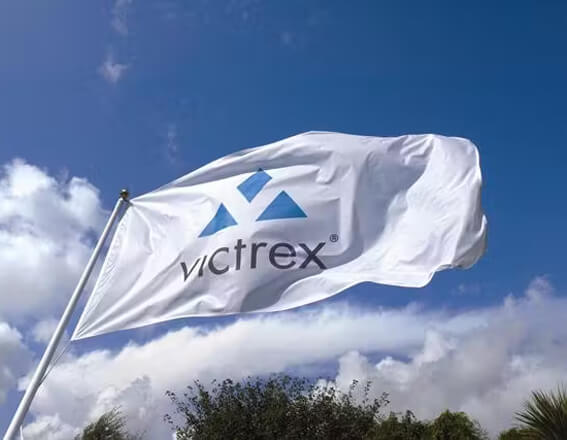 Weiße Victrex-Flagge weht im Wind vor blauem Himmel mit Wolken, symbolisiert Stärke und Innovation in der Fertigung.