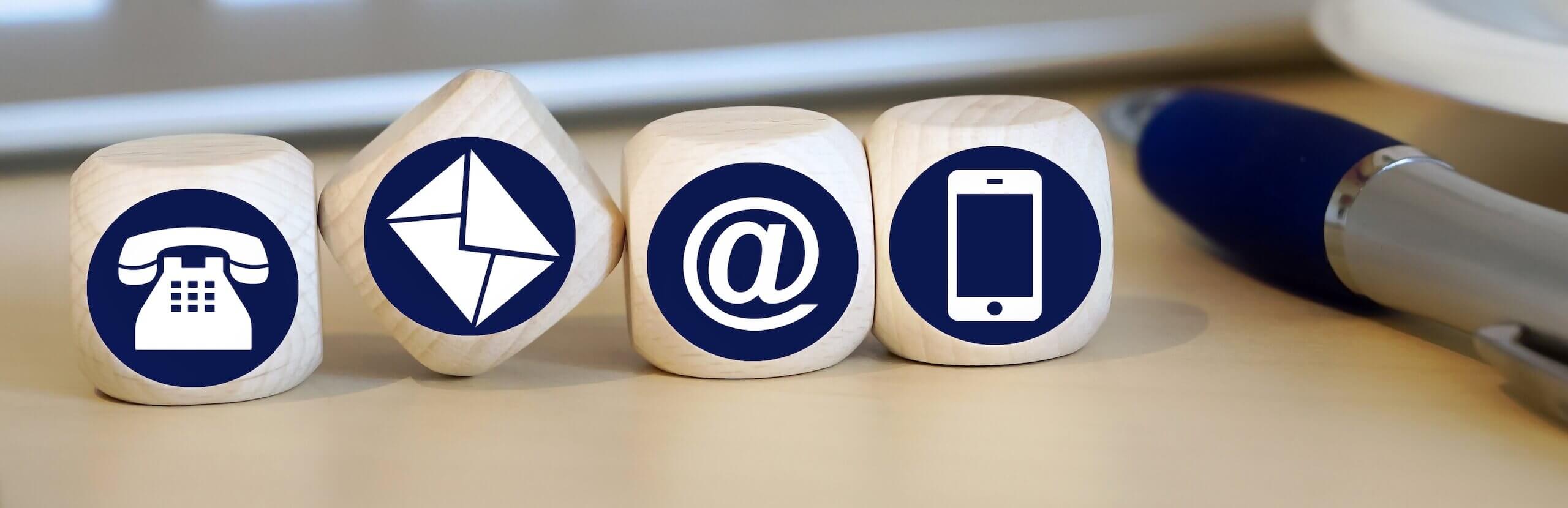 Holzwürfel mit Symbolen für Telefon, E-Mail, @-Zeichen und Smartphone auf einem Schreibtisch.